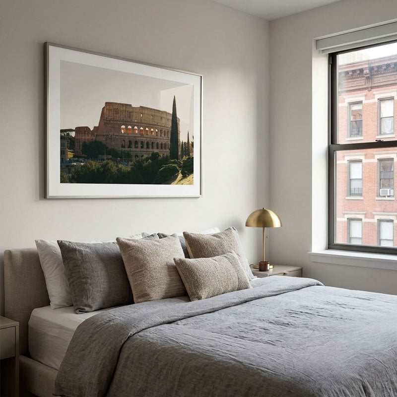 A framed art print of the Colosseum in Rome hangs on a light gray wall above a neatly made bed with gray and beige linens in a modern, softly lit bedroom.