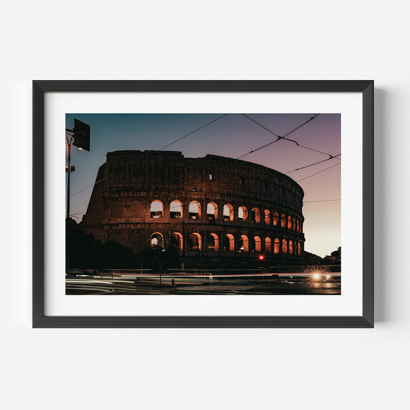 A framed photographic print of the Colosseum in Rome at dusk, shown against a light gray wall. The print features the ancient stone structure illuminated in warm orange light under a twilight sky, enclosed in a white mat and a black frame.