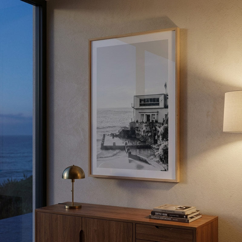 A framed black and white coastal photograph hanging on a textured beige wall above a wooden console table. The print is enclosed in a light wood frame with a white mat, shown in a styled interior with a lamp, books, and a window view of the ocean.