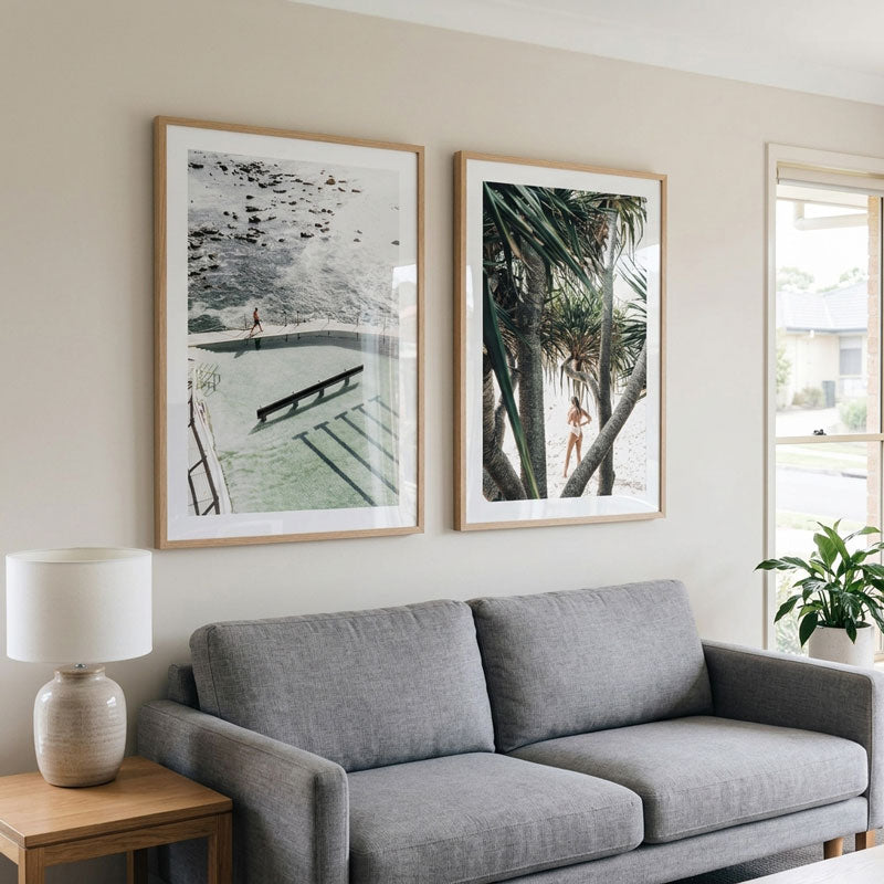 Two framed art prints hanging on a beige wall above a gray fabric sofa in a bright living room. The left print is a black-and-white architectural scene, and the right print shows a beach-themed scene with palm trees and a swimmer.