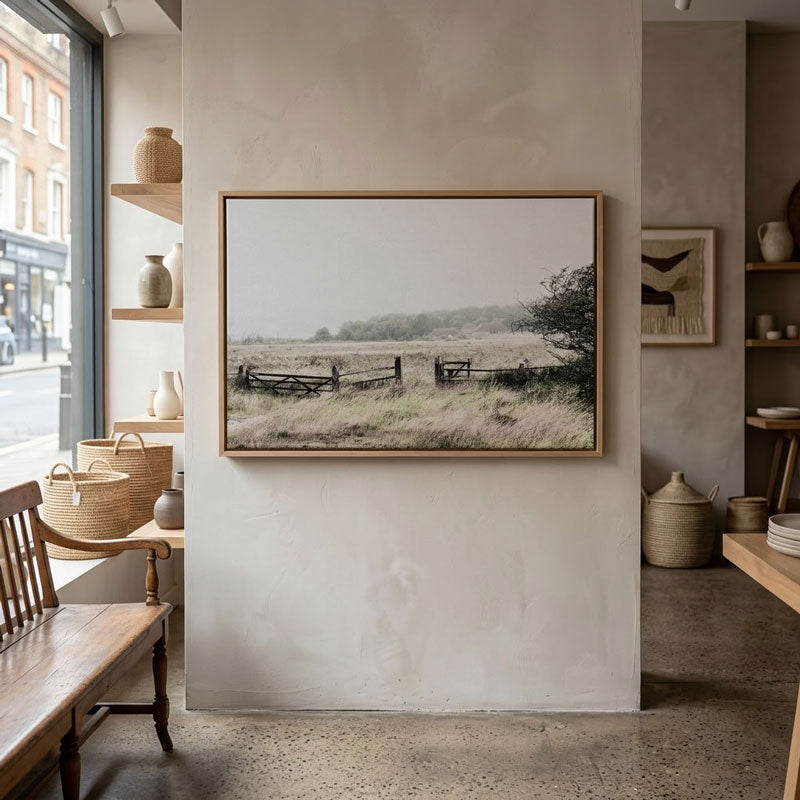 A framed landscape art print depicting a rustic field with a wooden fence, hung on a textured beige wall in a modern, rustic interior room.
