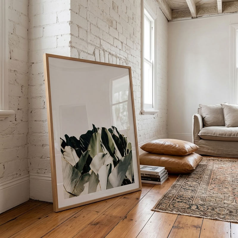 A large framed abstract botanical art print with green, white, and black leaves in a light wood frame, leaning against a white brick wall in a bright, modern living room with a brown leather sofa and wooden floors.