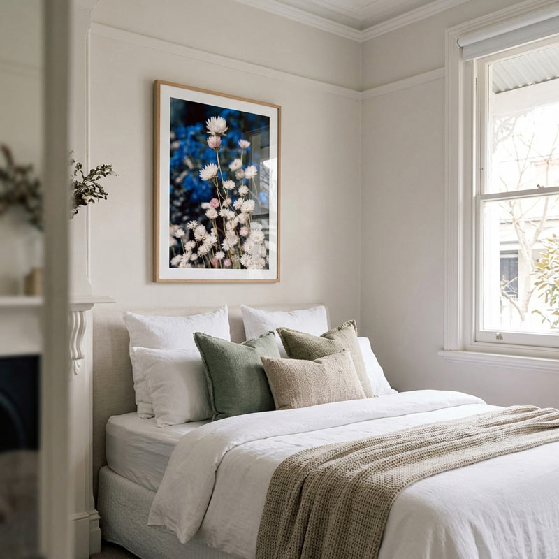 A framed floral art print hangs on a light beige wall above a neatly made bed with white linens, sage green pillows, and a textured beige throw blanket in a bright, modern bedroom.