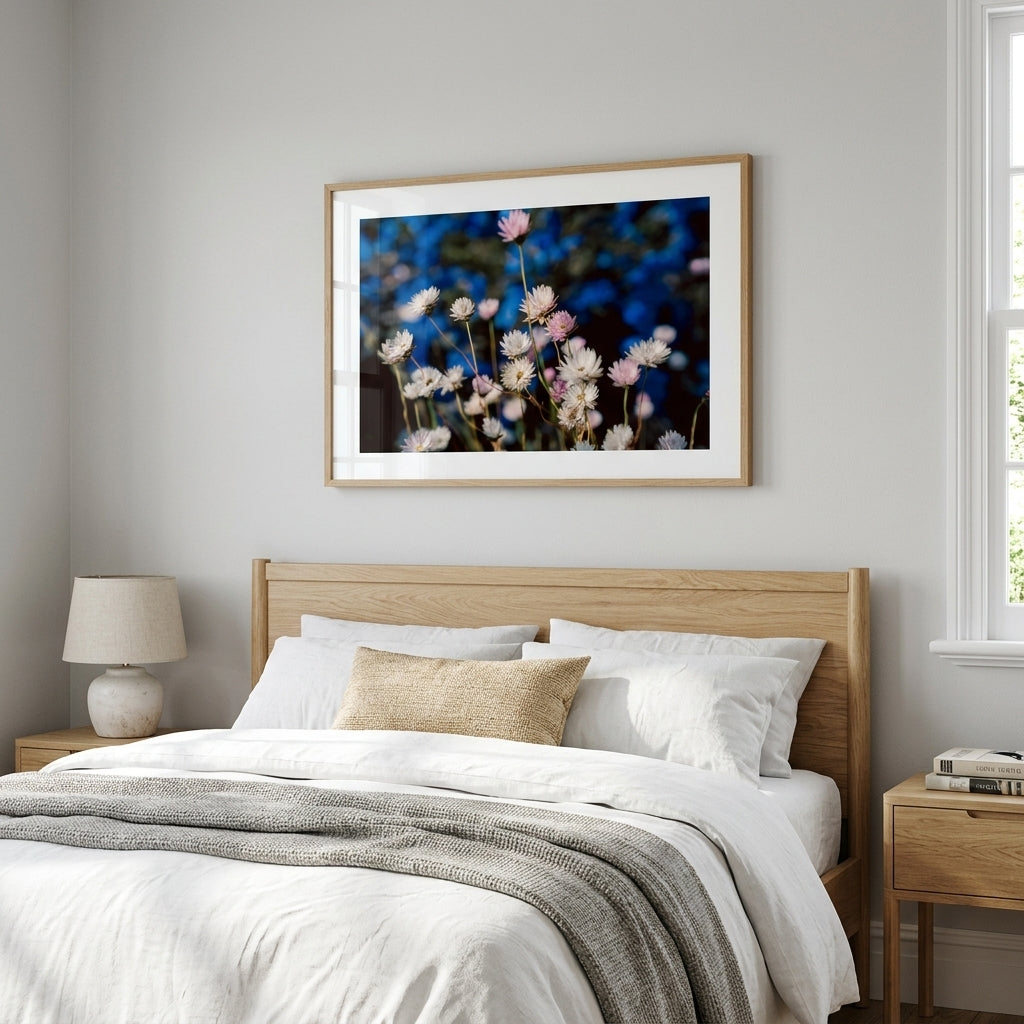 A framed floral art print hangs above a neatly made bed in a bright, modern bedroom. The print features white and pink flowers against a dark blue background, enclosed in a light wood frame with a white mat.