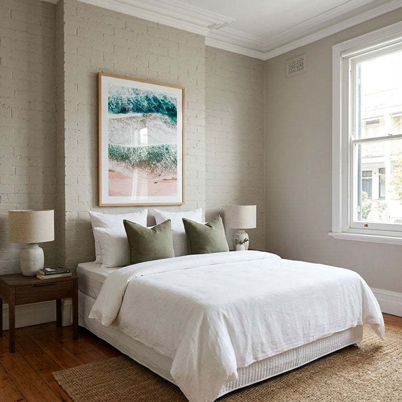 A styled bedroom interior featuring a neatly made bed with white linens and olive green pillows, set against a light brick wall with a large framed wave artwork. The room includes wood flooring, bedside tables with white lamps, and a window on the right.