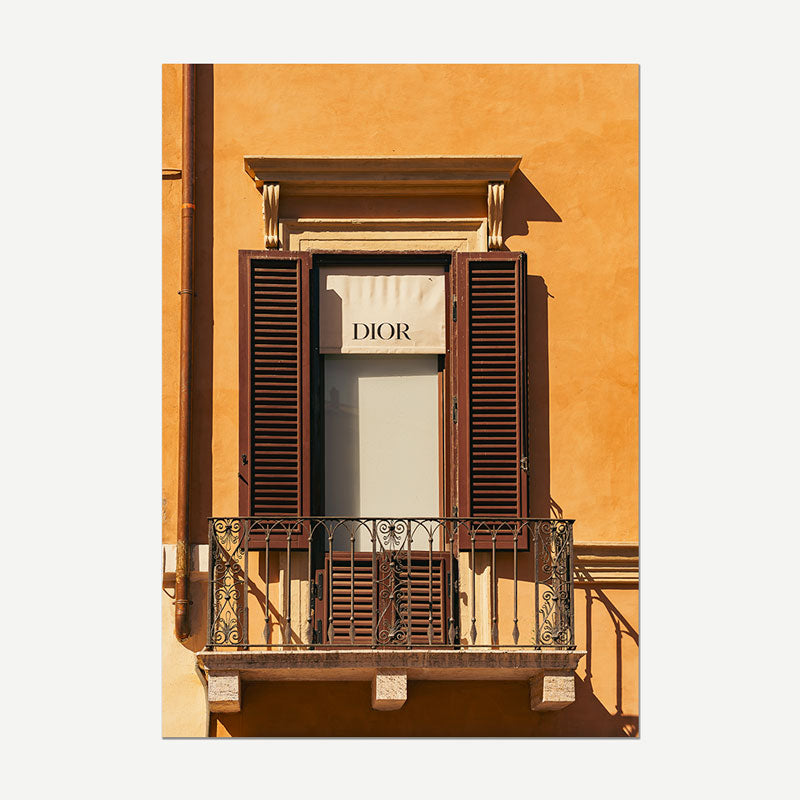 A vertical art print depicting a classic European building facade with a warm ochre-orange stucco wall, dark brown wooden shutters, and a small balcony. A white banner on the window displays the brand name 'DIOR'.