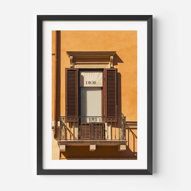 A framed art print featuring a photograph of a classic building facade with a window, dark brown shutters, and a Dior sign, presented in a black frame with a white mat against a light gray wall.