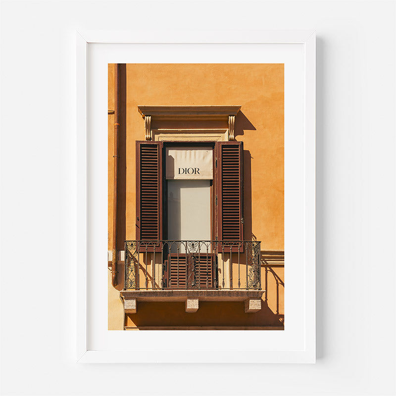 A framed art print featuring a photograph of a building facade with a window, dark brown shutters, and a Dior sign, presented in a white frame against a light gray background.