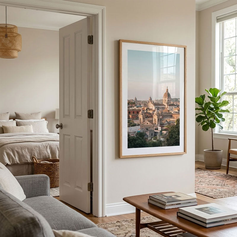 A large framed art print depicting a scenic city view with domes, hung on a beige wall in a modern, well-lit living space with a sofa, coffee table, and potted plant.