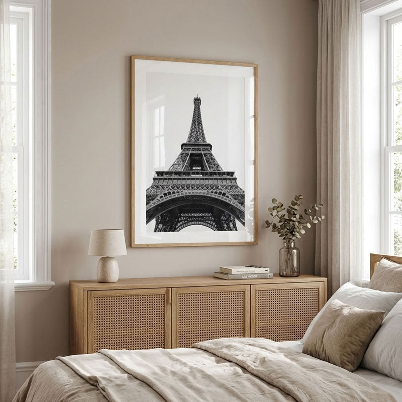 A framed black and white art print of the Eiffel Tower hanging on a beige wall above a wooden nightstand in a styled bedroom setting.