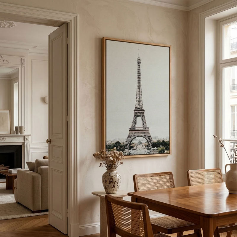 A large framed vertical art print of the Eiffel Tower hanging on a beige wall in a styled dining room with a wooden table, woven chairs, and a vase.