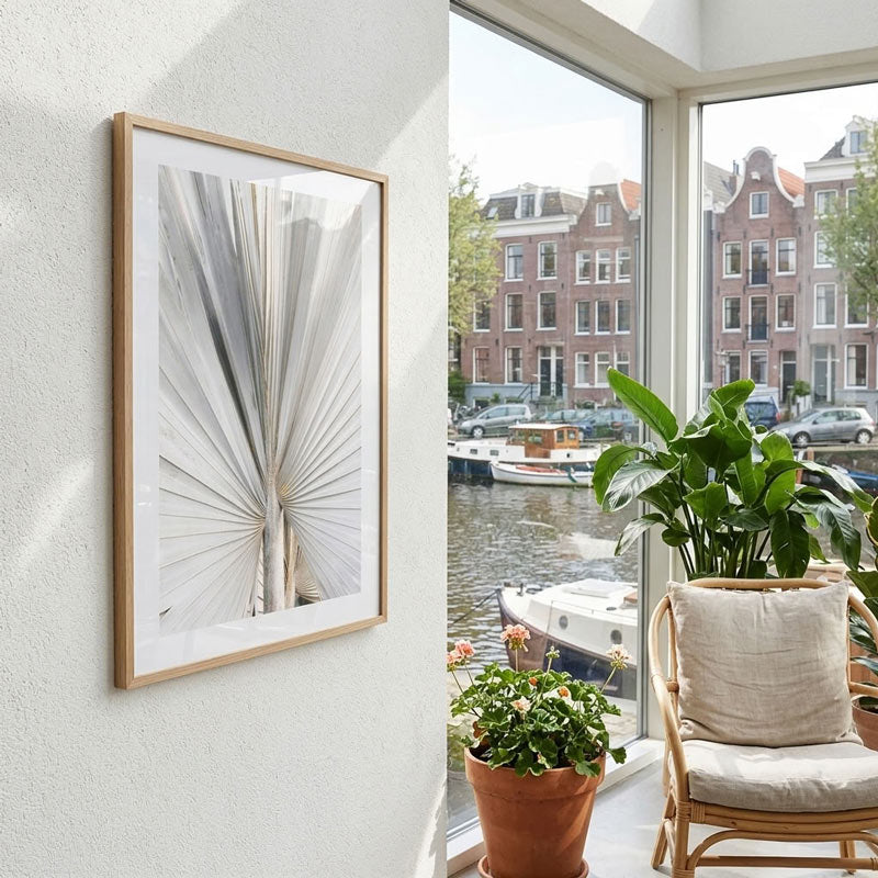 A framed botanical art print with a light wood frame hangs on a white wall next to a large window overlooking a canal with boats and Amsterdam-style buildings. The room includes a potted green plant and a woven chair with a beige cushion.