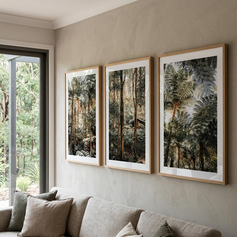 Three framed forest art prints with light wood frames and white mats are displayed on a textured beige wall above a gray sofa, with a window showing green foliage outside.