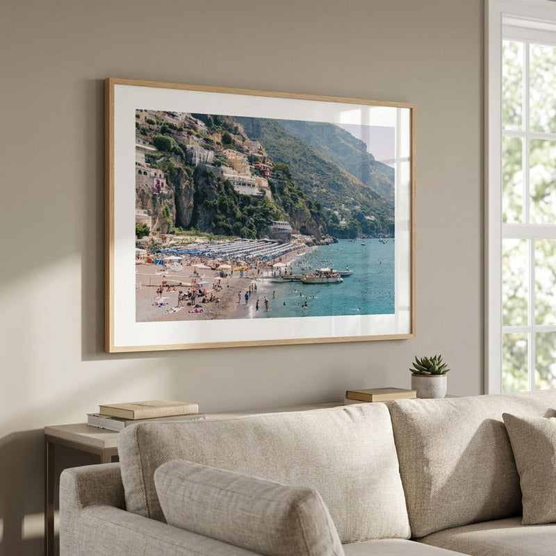 A framed coastal beach print hangs on a beige wall above a light beige sofa in a styled living room. The artwork shows a sunny seaside town with blue water, sandy beach, and green hillsides.
