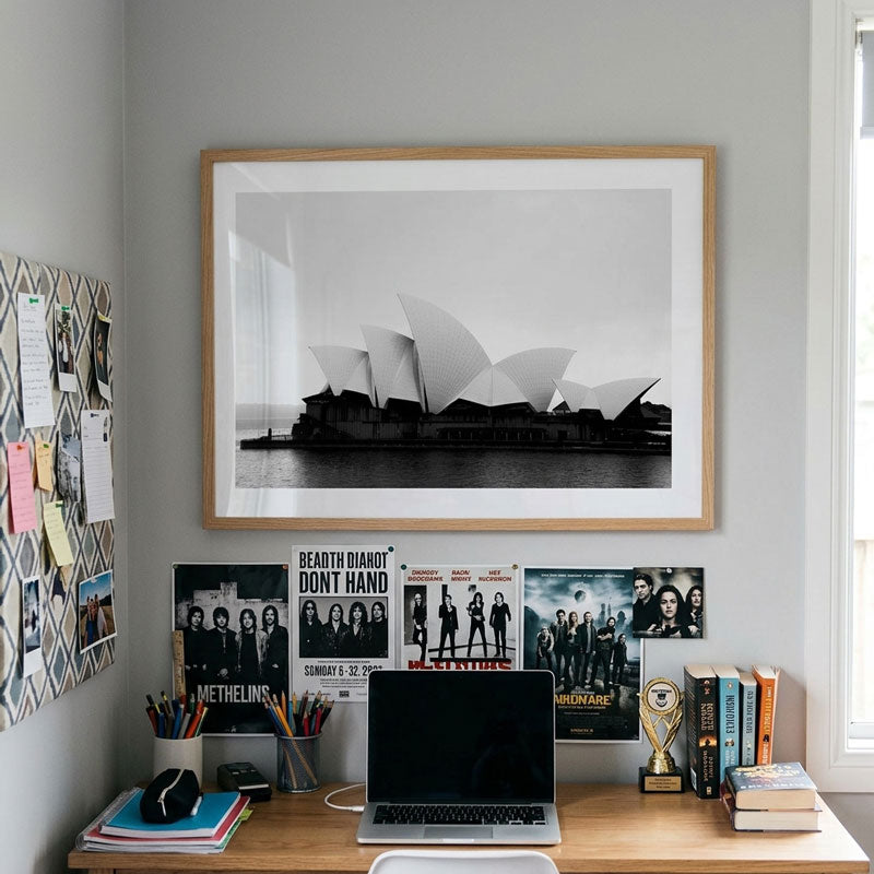 A framed black and white photograph of the Sydney Opera House hanging on a light gray wall above a wooden desk with a laptop, books, and posters.