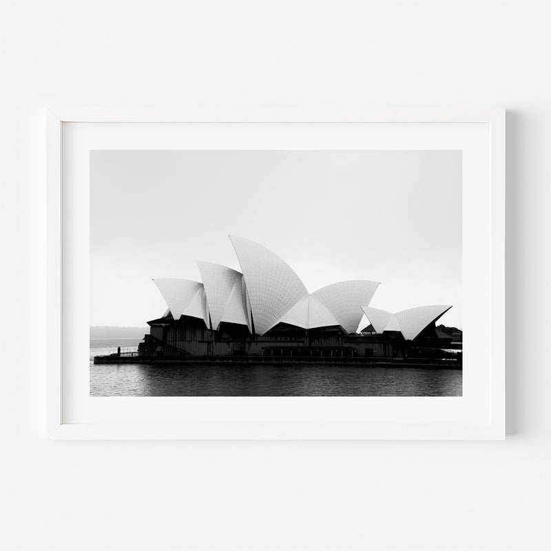 A framed black and white photograph of the Sydney Opera House, shown in a white frame against a clean white wall.