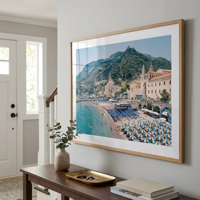 A large framed beach and coastal town art print hangs on a light gray wall above a wooden console table in a styled interior. The print depicts a sunny seaside town with blue umbrellas, turquoise water, mountains, and buildings, surrounded by a white mat and light wood frame.