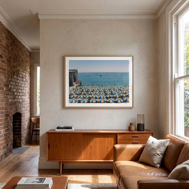 A framed square art print depicting a crowded beach filled with colorful umbrellas under a blue sky, displayed on a beige wall in a modern living room setting with a wooden credenza and leather sofa.