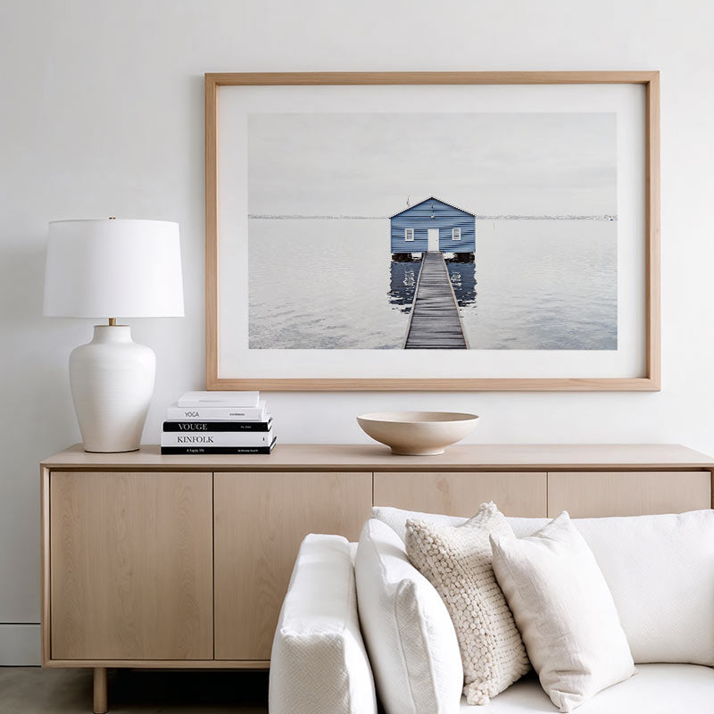 Framed artwork of a blue house on a pier above a wooden sideboard with decor items.