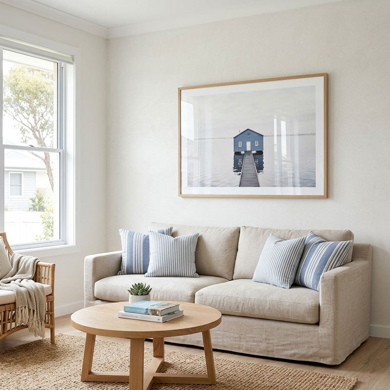 A beige fabric sofa in a bright, modern living room, styled with blue and white striped pillows, a round wooden coffee table, and a large framed art print on the wall depicting a blue boat on a pier.
