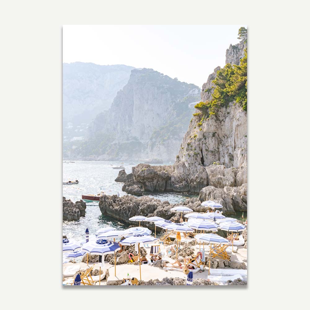 La Fontelina Beach Club in Capri with blue and white beach umbrellas and sun desks on a rocky Mediterranean shore, bathed in a golden late afternoon light, evoking coastal elegance and tranquil Italian summer vibes.
