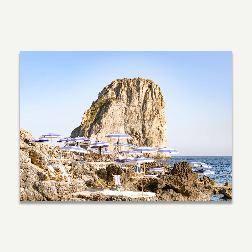 Late afternoon photograph of La Fontelina Beach Club in Capri, featuring blue and white umbrellas on rocky shores with Faraglioni rocks in the backdrop, evoking Mediterranean elegance and coastal charm.