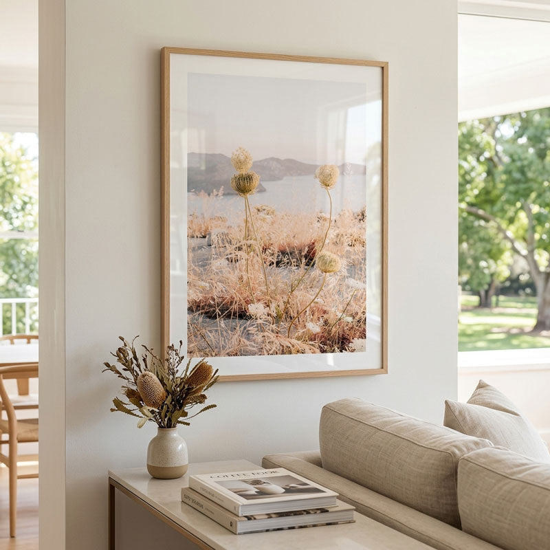 A large framed art print depicting a dry grass field with pampas grass heads and distant hills under a soft sky, styled in a modern living room above a beige sofa.