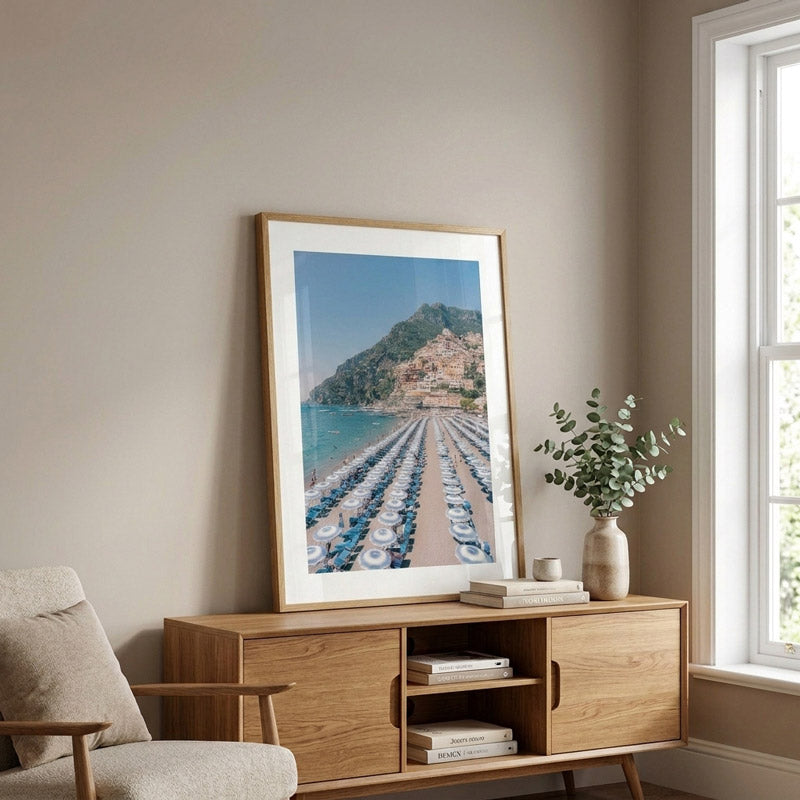A large vertical framed art print depicting a scenic coastal town with rows of beach umbrellas by the sea, styled on a light wood sideboard in a modern living room setting.