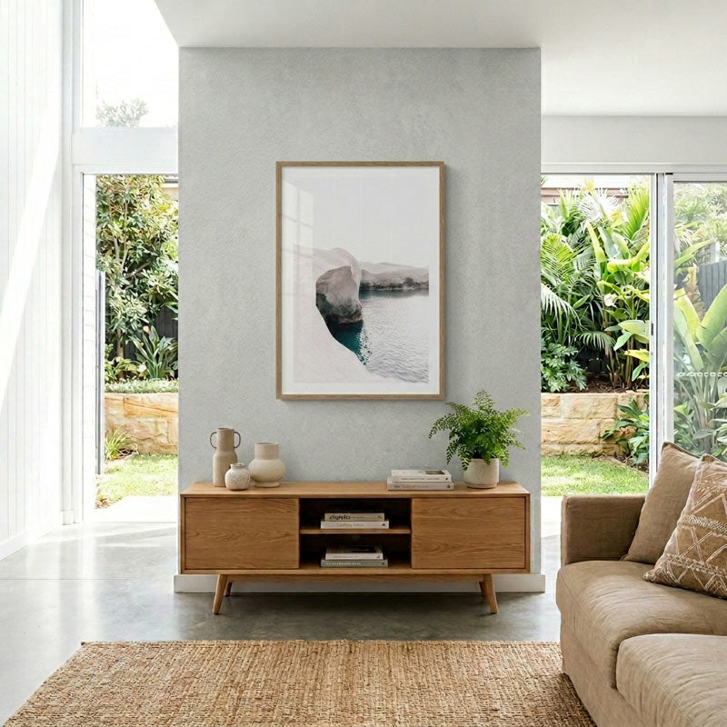 A framed abstract landscape art print hangs on a textured light gray wall above a wooden media console in a modern living room. The print depicts a coastal scene with white surf, blue water, and distant rocky mountains.