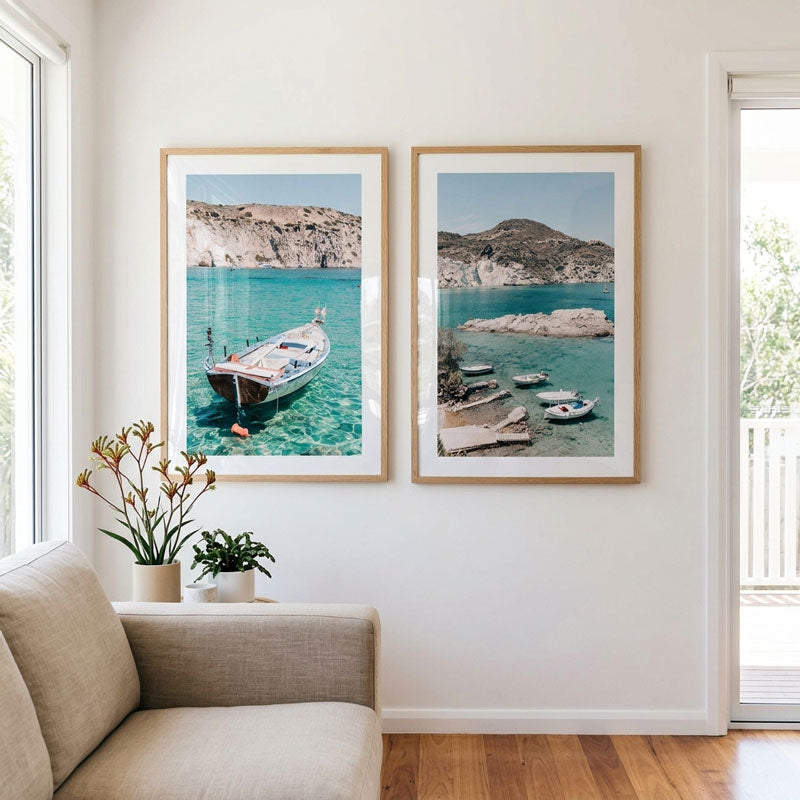 Two framed coastal art prints with turquoise water and boats hang on a white wall above a beige sofa in a bright living room.