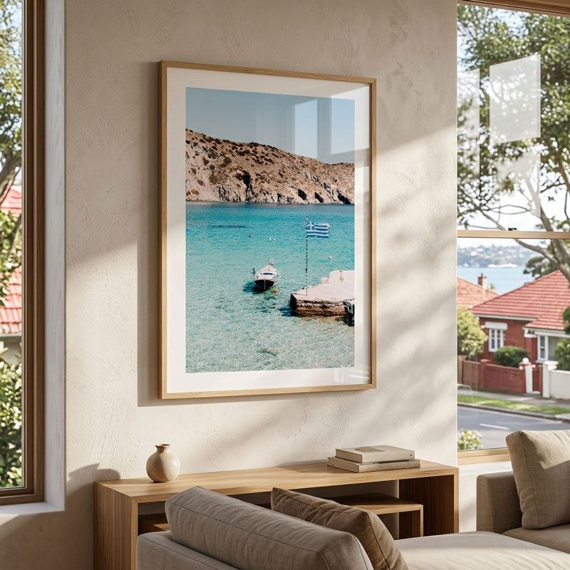 A framed vertical art print of a Greek coastal bay with turquoise water, rocky cliffs, and a small boat, displayed on a beige wall above a wooden console table in a modern living room setting.