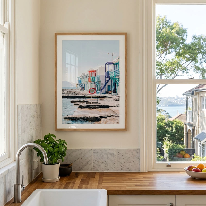 A framed art print depicting a colorful seaside pier and harbor scene hangs on a cream kitchen wall above a wooden countertop with a sink, potted plant, and fruit bowl. The print is shown in a real interior environment, so this is Lifestyle rather than Scenery or Product/Packaging.