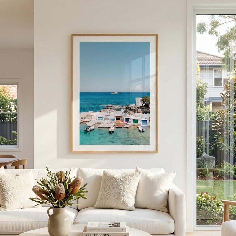 A framed art print depicting a scenic coastal harbor with turquoise water and white buildings, hanging on a cream wall above a white sofa in a bright, modern living room.