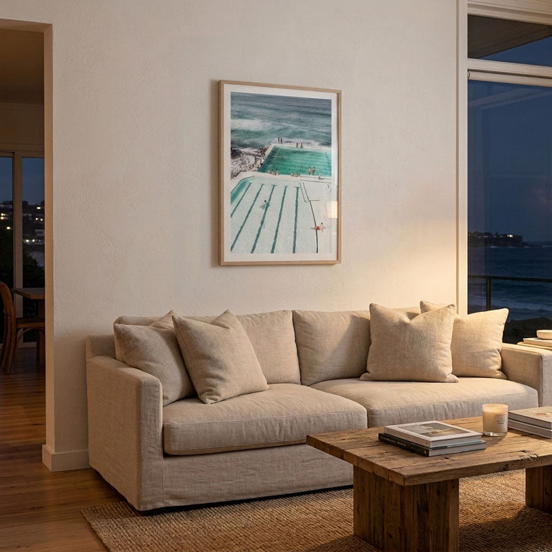 A framed art print of an outdoor swimming pool and beach scene hangs on a beige wall above a light beige sofa in a modern living room setting.