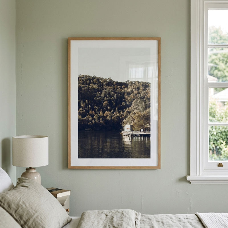 A framed landscape art print of a forested hillside and lake with a small dockhouse, displayed on a sage green bedroom wall above a bed.