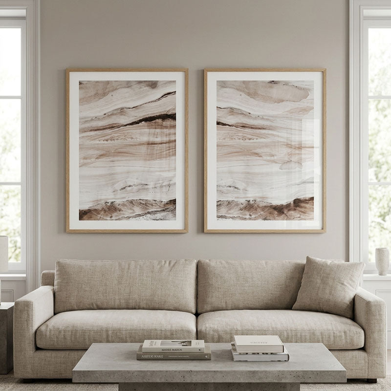 Two framed abstract art prints with soft beige, brown, and white brushstrokes hang on a light gray wall above a beige sofa in a bright living room.