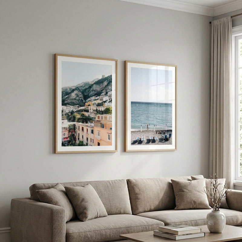 Two framed coastal landscape prints hang on a light gray wall above a beige sofa in a styled living room. The left print shows a hillside town with mountains, and the right print shows a seaside beach and ocean.