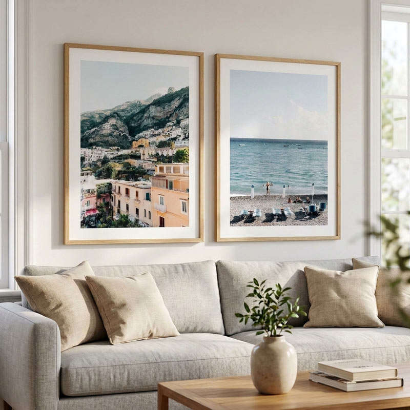 Two framed coastal and mountain prints displayed side by side above a light gray sofa in a bright living room. The left print shows a hillside town with terracotta buildings, and the right print shows a seaside beach with blue water and lounge chairs.