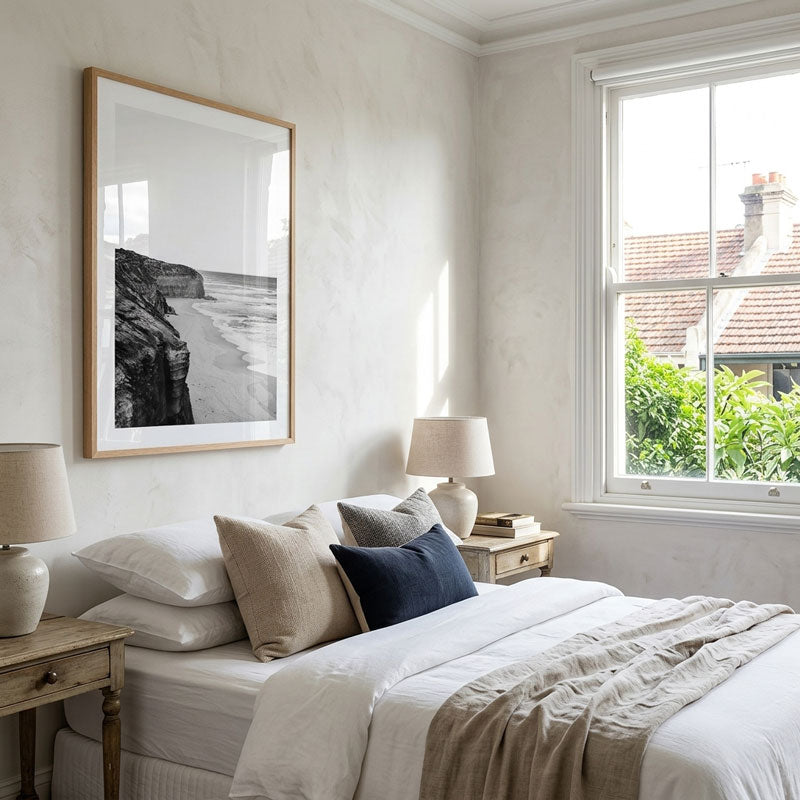A styled bedroom interior with a bed dressed in white and beige linens, multiple pillows including a dark navy accent, bedside tables with lamps, and a large framed black-and-white coastal print on the wall. A window with white trim shows greenery and rooftops outside.