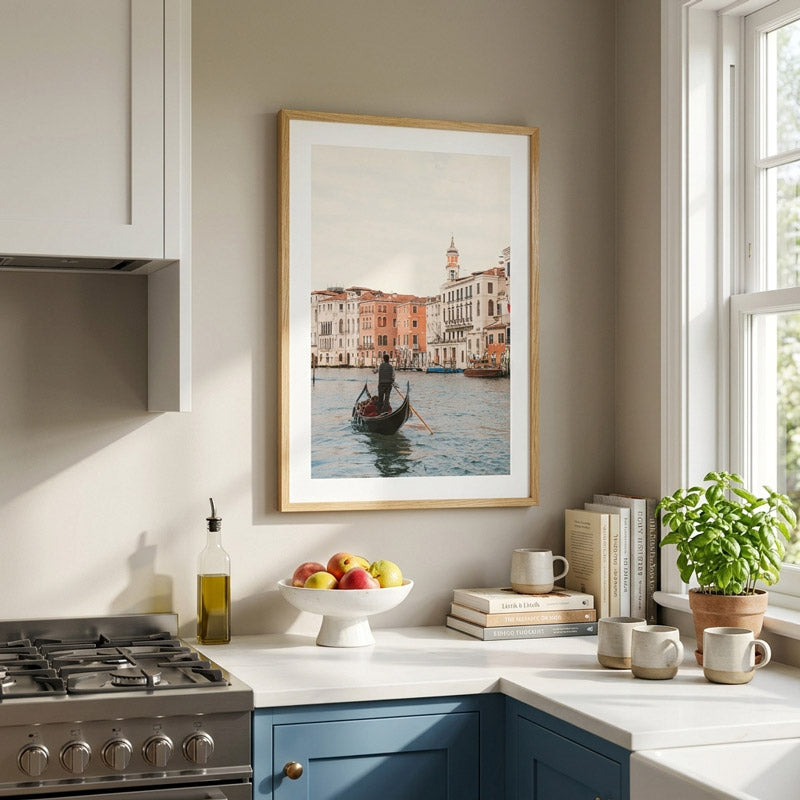 A framed art print depicting a gondola on the Grand Canal in Venice hangs on a beige wall above a kitchen counter with blue cabinets, a bowl of fruit, books, mugs, and a potted plant.