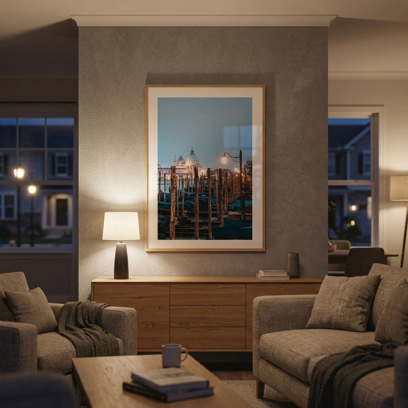 A framed vertical art print depicting a waterfront pier at dusk, displayed on a textured gray wall in a modern, warmly lit living room setting.