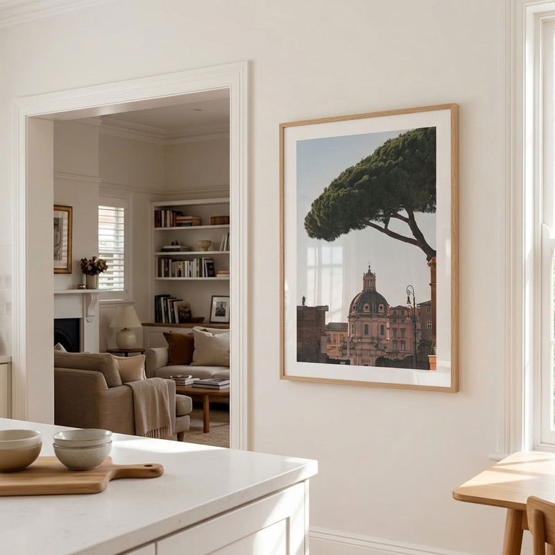 A framed art print of a Tuscan landscape with a prominent green tree and a domed building, displayed on a cream-colored wall in a bright, modern kitchen/living room setting.
