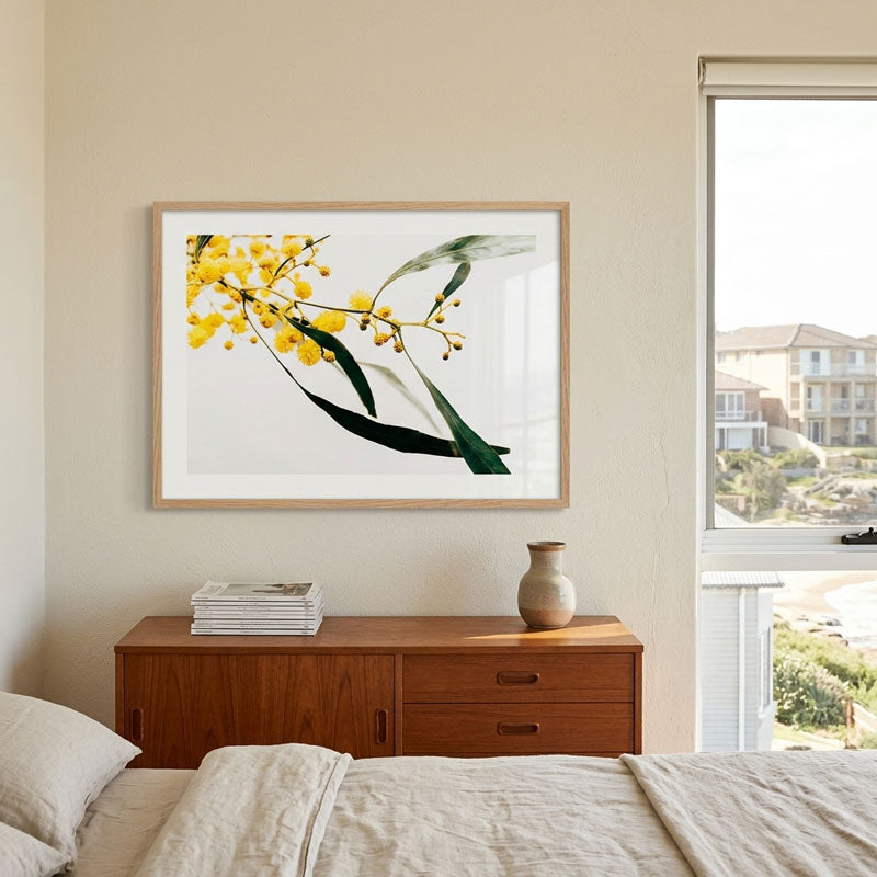 A framed botanical art print of yellow mimosa flowers and green leaves, displayed on a beige wall above a wooden nightstand in a modern bedroom setting.