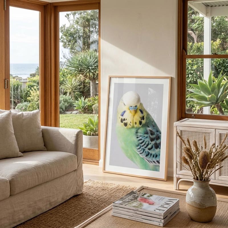 A framed art print of a green and blue budgerigar parrot leaning against a wall in a modern, sunlit living room with a beige sofa and large windows overlooking a garden and ocean.
