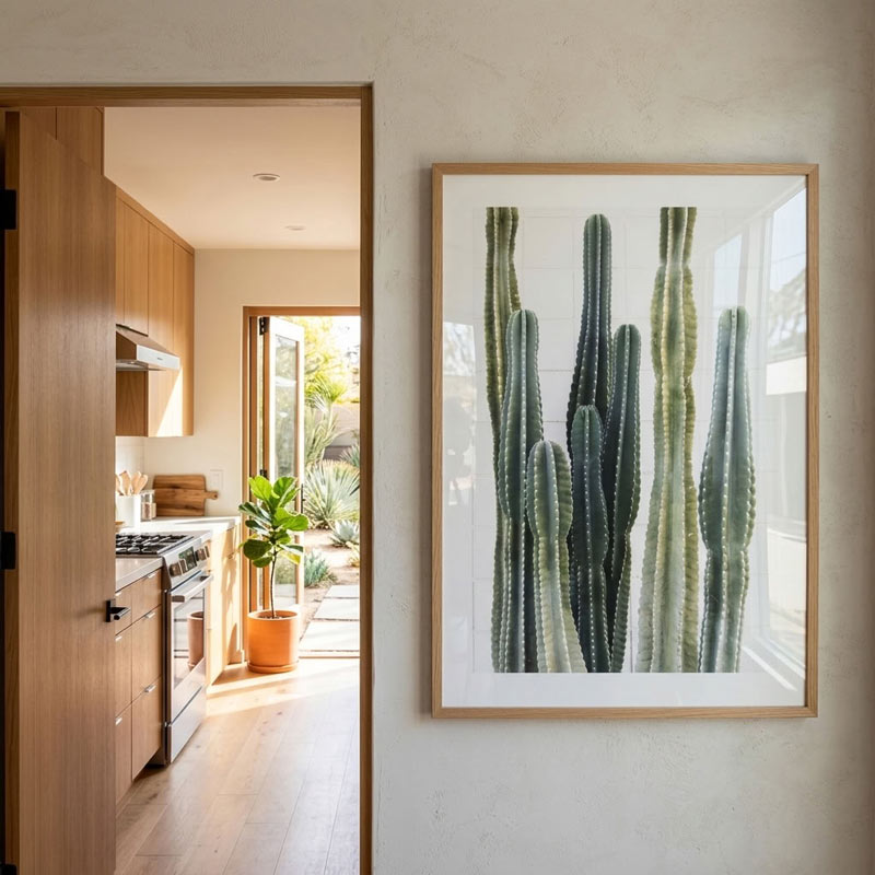 A framed botanical art print of green cacti on a white background, hung on a textured beige wall next to an open doorway revealing a modern kitchen and patio.