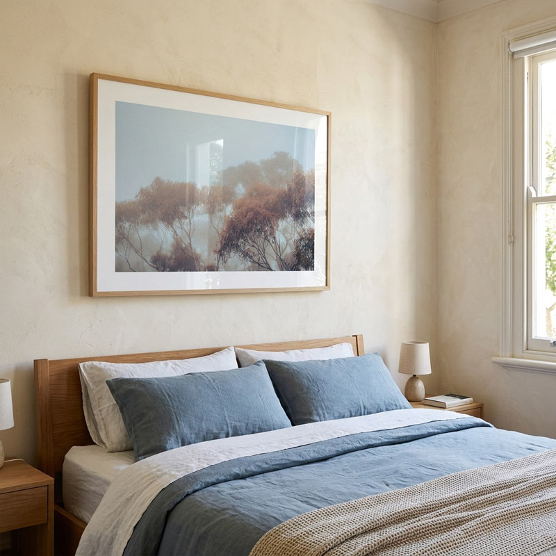 A framed landscape art print hangs on a textured beige wall above a neatly made bed with blue and white linens in a softly lit bedroom.
