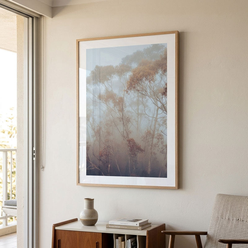 A framed art print depicting a misty forest with soft blue-gray and brown trees, hung on a beige wall above a wooden cabinet in a modern, sunlit room.