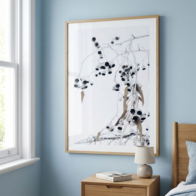 A framed botanical art print featuring black and brown botanical elements on a white background, hung on a light blue wall above a wooden nightstand in a bedroom setting.