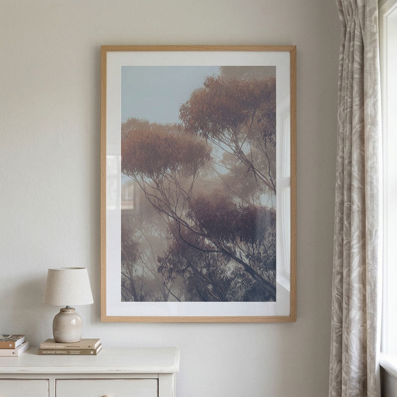 A framed art print depicting a misty forest of trees with soft brown and purple foliage, hung on a light gray wall above a white sideboard with a lamp and books, next to a patterned curtain.