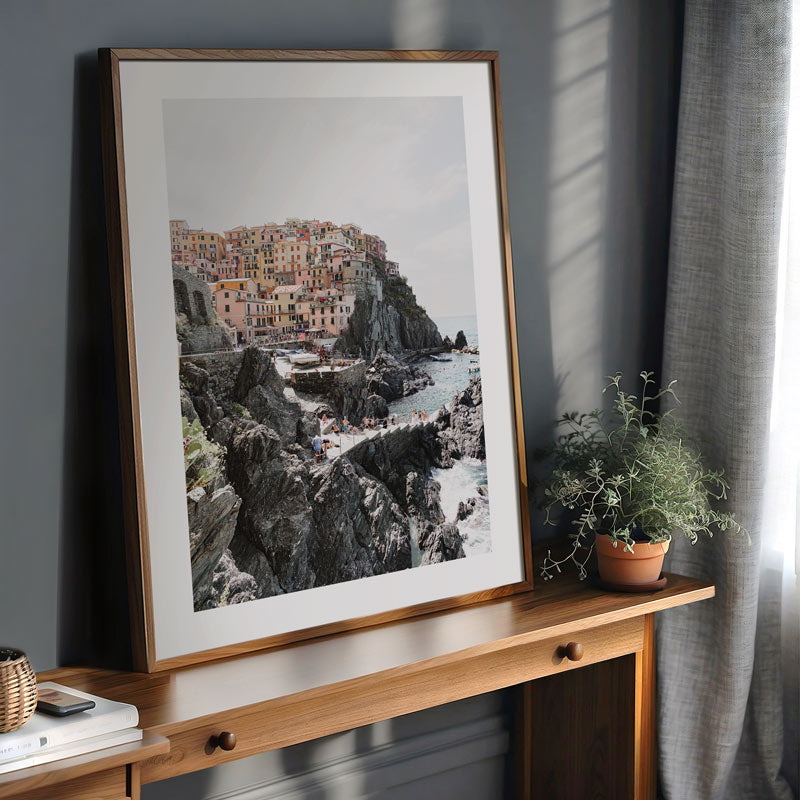Framed photograph of a coastal village on a wooden shelf with plants and books.